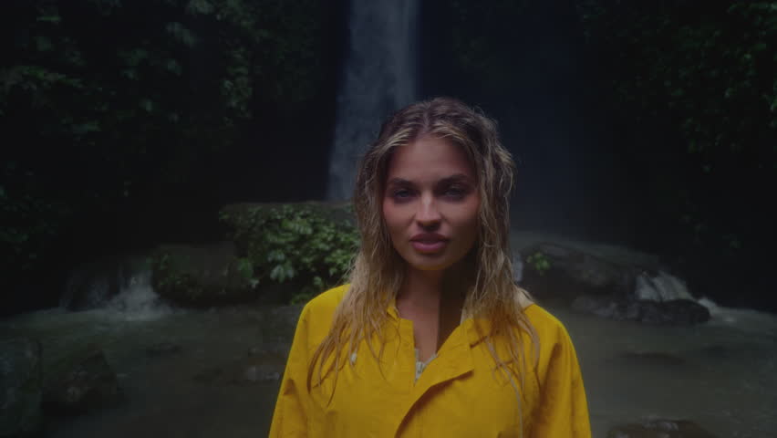 Portrait: beautiful blonde Slavic girl exploring a waterfall in Bali Indonesia. Woman in a yellow raincoat with beauty face in tropical jungle is enjoying vacation, trip. Rainy season, water lake