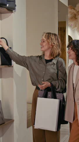 Vertical view of joyful young woman choosing new bag and asking girl friend for advice while shopping together