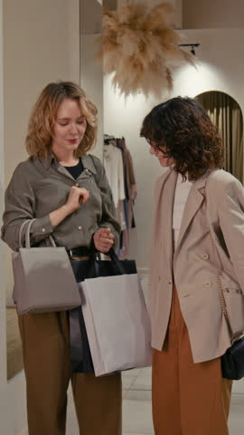 Vertical view of joyful young woman choosing new bag and asking girl friend for advice while shopping together
