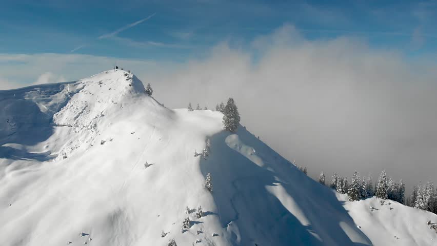Aerial flyover over a mountain peak with hikers overlooking a cloud inversion above alpine slopes with trees covered in fresh snow powder with Swiss Alps in background on a sunlit winter day