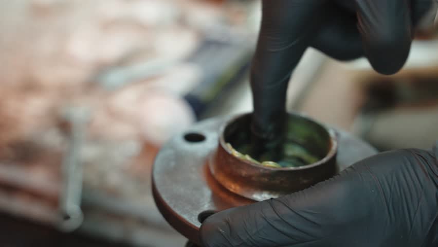 Car service worker wearing black gloves carefully applies grease to a metal wheel bearing, ensuring smooth operation and preventing wear and tear