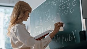 Teacher writing math equations on chalkboard looking book closeup. Woman educator solving mathematical tasks on blackboard classroom. Blonde female tutor in glasses teaching algebra in high school. - Powered by Shutterstock - Get 15% off with code: PIKWIZARD15