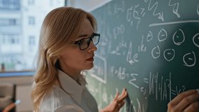 Algebra educator writing on chalkboard explaining mathematical formulas in school class closeup. Serious woman teacher in glasses teaching math in college classroom. Female university professor work. - Powered by Shutterstock - Get 15% off with code: PIKWIZARD15