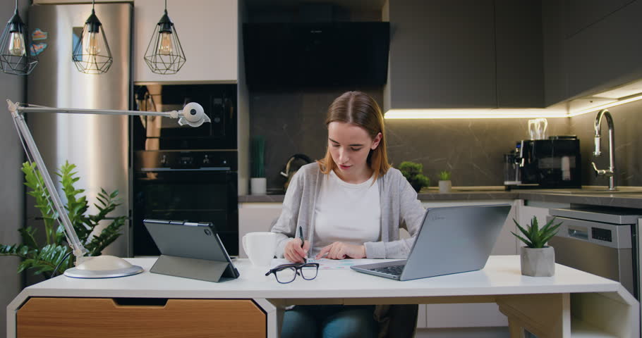 Online from home watching web class lesson or listening tutorial. Woman entrepreneur using laptop writing notes in modern at home office desk. Distance education concept.