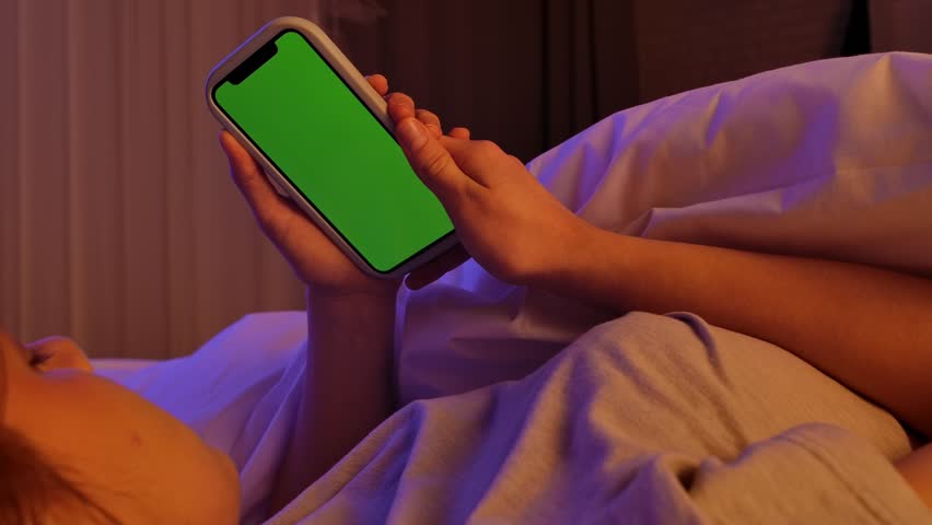 Young woman laying in bed on the pillow covered with blanket in pink neon light, holding smartphone with chroma key green screen background in hand. Slow motion.