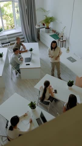Man with long hair conducts business training in big audience. Professional coach talks to people sitting at white desks upper angle view slow motion Vertical Shot.