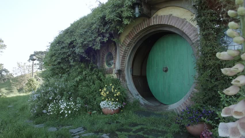 A lush hobbit house with a green round door, brick archway, climbing vines, and colorful flowers, set in grassy hills under soft daylight in Hobbiton, New Zealand.