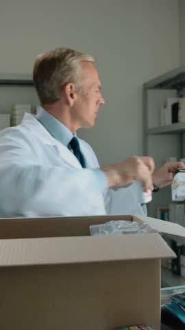 Pharmacist unpacking delivery from stock indoor of drug store interior. Caucasian mature man fills the shelves with medicine pills, ampoules of saline etc. Middle view, daylight. Vertical shot.