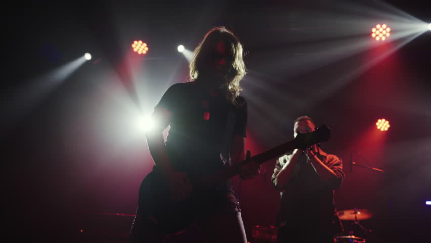 Female guitarist playing electric guitar on stage with a singer in the background, illuminated by stage lights in slow motion