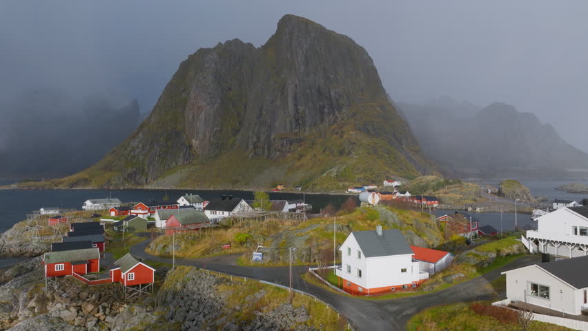 Aerial view of Hamnoy village and Hamnoy bridge with charming red cabins, and the towering Lofoten peaks under a moody, overcast sky