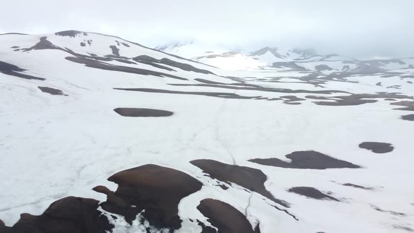 Aerial drone footage rising up and revealing dark black and brown volcanic Iceland mountain landscapes covered in white snow during a cloudy and misty day. background is very cloudy and foggy.