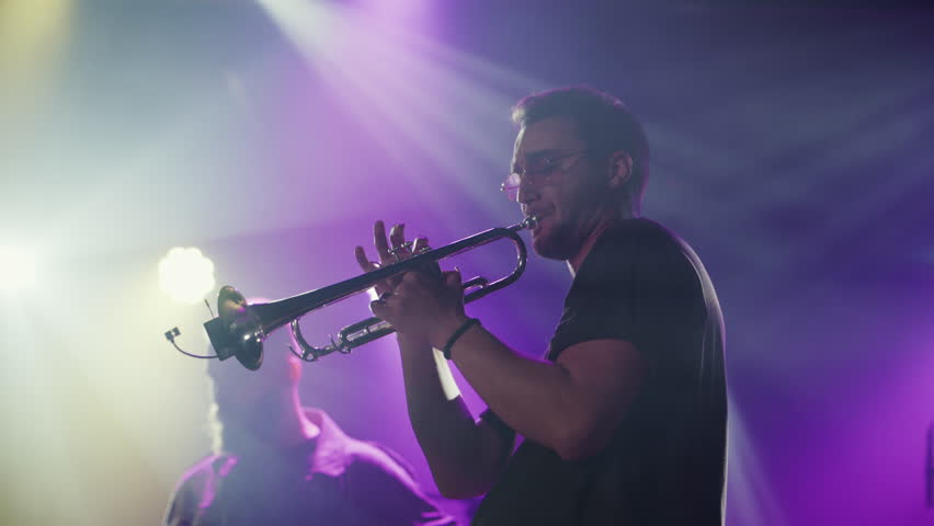 Skilled trumpet player delivering passionate performance under vibrant purple stage lights, revealing musical artistry through slow motion footage