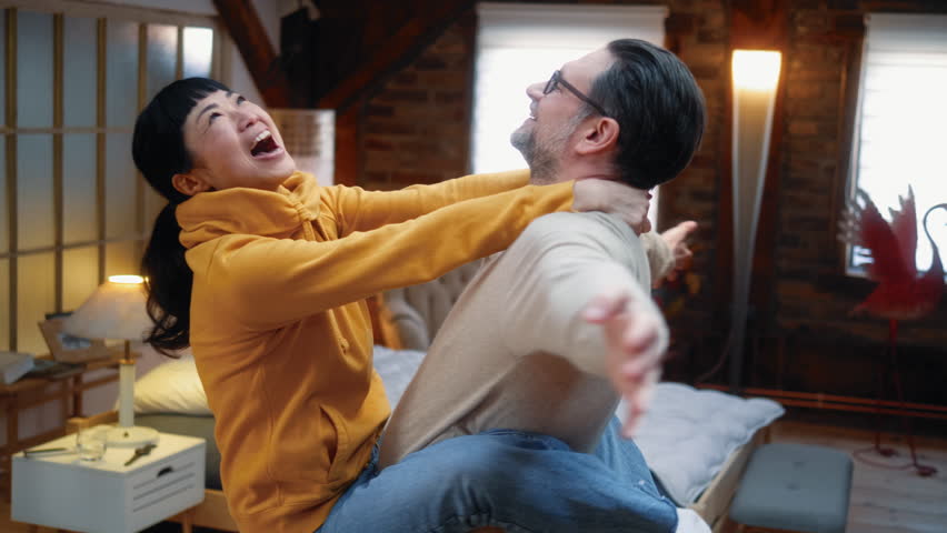 White man holding Japanese woman in his arms dancing and twirling cozy bedroom, excited Asian wife laughing hugging Caucasian husband feeling happy having fun together at home.