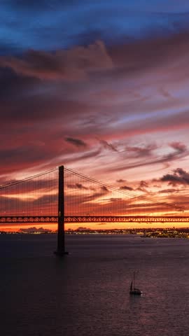 Time-lapse of 25 de Abril Bridge famous tourist landmark of Lisbon connecting Lisboa and Almada on Setubal Peninsula over Tagus river in the evening twilight. Lisbon, Portugal. Camera pan effect