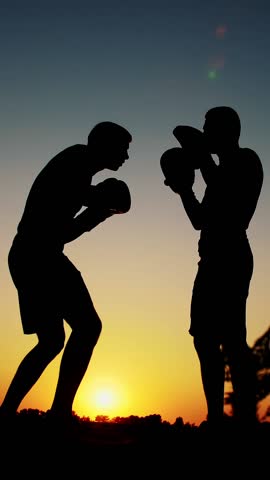 Two dark male figures, at sunrise, against the light, boxing, fighting in sparring, training in a pair of techniques of strikes, near water, in summer