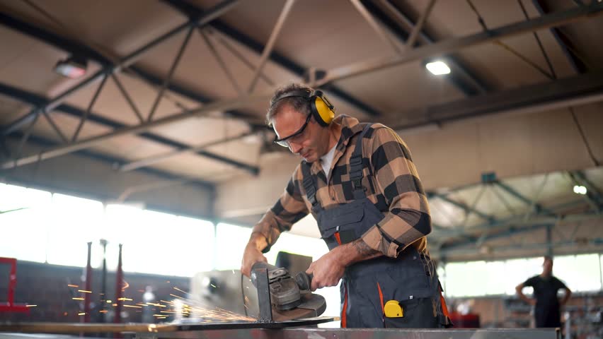 Industrial worker wearing safety glasses and ear protection using angle grinder to cut metal in a workshop