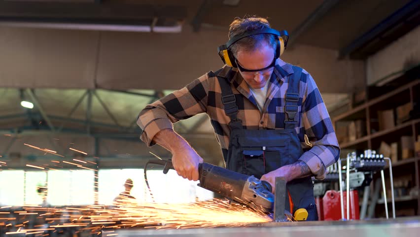 Skilled metalworker wearing safety glasses and ear protection uses an angle grinder, creating sparks while cutting metal