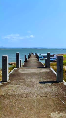 A concrete pier extending out into a lake with turquoise water. There are several boats moored at the edge of the water and several people are seen at the end of the pier.