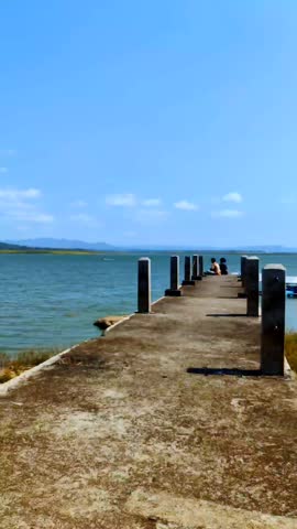 A concrete pier extending out into a lake with turquoise water. There are several boats moored at the edge of the water and several people are seen at the end of the pier.