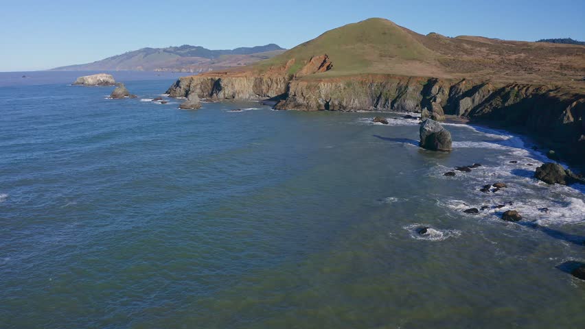 Breathtaking aerial view of the rugged California coastline along Highway 1. Steep cliffs covered in green vegetation rise above the deep blue ocean, with waves crashing against rocky formations. The 