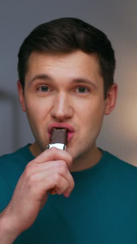 Portrait of Adult Man Eating Sweet Food and Looking at Camera. Real Person on Diet Consuming Chocolate Candy Bar as Lunch Snack. Confident Male Human Inside Cozy Living Room Alone with Sugary Fastfood