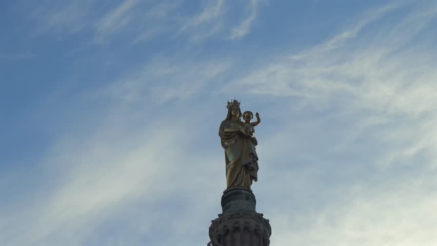Close-up shot of the golden Madonna and Child statue atop Notre-Dame de la Garde in Marseille. The camera tilts upward against a dynamic blue sky with wispy clouds.