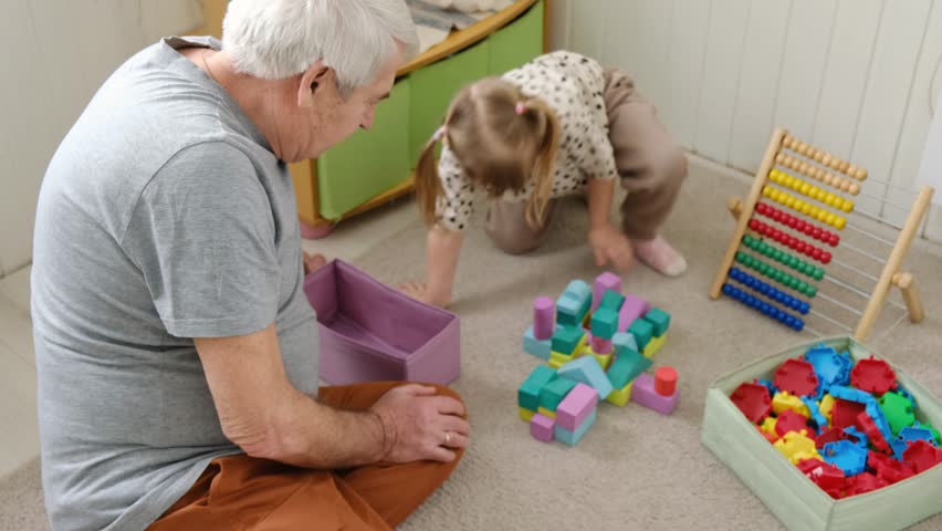 Grandfather playing with little baby. Caring granddad and cute toddler grandchild sitting on carpet floor together, having fun at home. Happy old senior man teach granddaughter playing colorful blocks