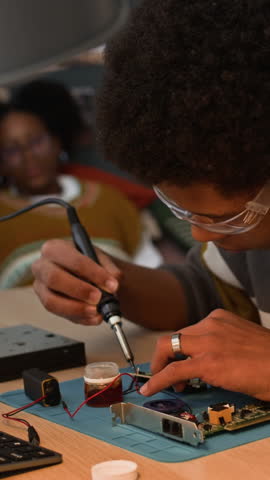 Vertical of concentrated African American teenage boy wearing protective goggles soldering chips with soldering iron on table in his room with his girlfriend watching him working