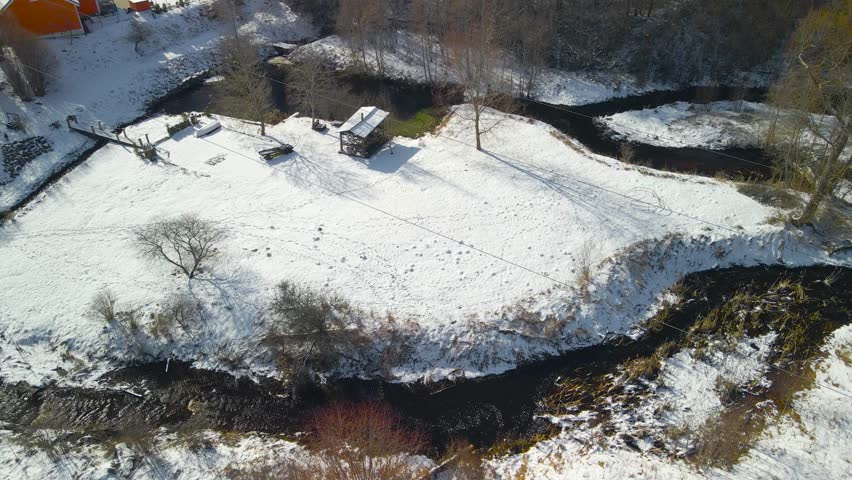 Aerial drone footage of small dark narrow rivers or water streams flowing in winter white snow covered forest during a sunny day with some houses visible in the back. Rivers turn and twist on land.