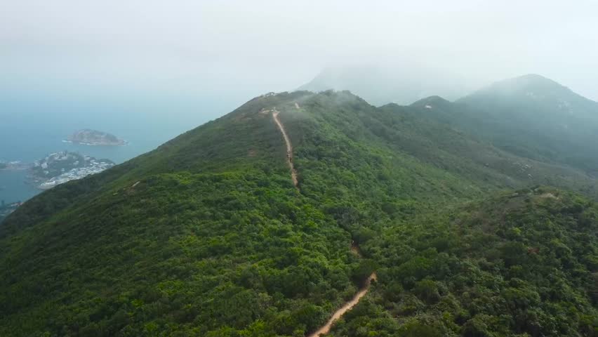 Aerial drone footage flying towards and closer to a large green louscious mountain during a cloudy day with a muddy brown dirt road leading up the hillside. Large blue ocean is visible behind the hill