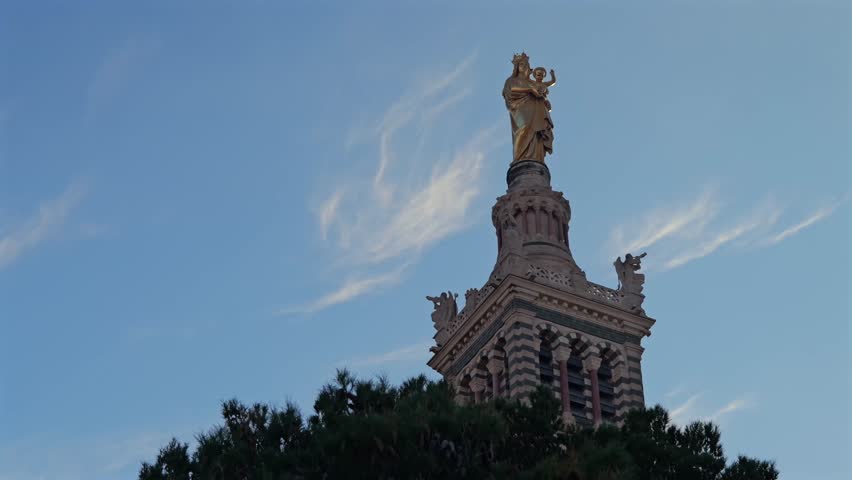 The golden Madonna and Child statue stands atop Notre-Dame de la Garde in Marseille. Captured from a low angle, the intricate architecture contrasts with the blue sky and wispy clouds.