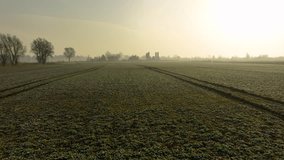 Drone shot of an open field at sunrise with distant farm buildings, frost-covered grass, and tractor tire tracks leading into the misty horizon - Powered by Shutterstock - Get 15% off with code: PIKWIZARD15