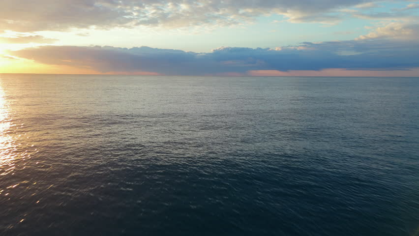 Drone glides low over the ocean at sunrise, capturing golden reflections on the calm water. The horizon blends with vibrant clouds, creating a peaceful seascape