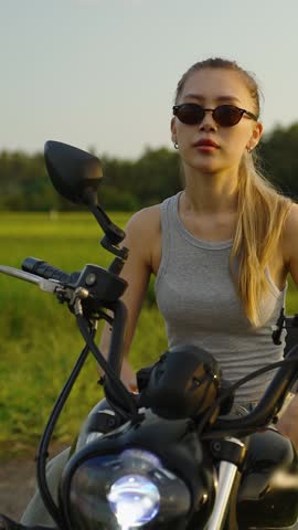 Confident young woman wearing sunglasses leans on a motorcycle, moto driver in a green rural landscape at sunset. Bike driving and moto trip concept. Vertical shot