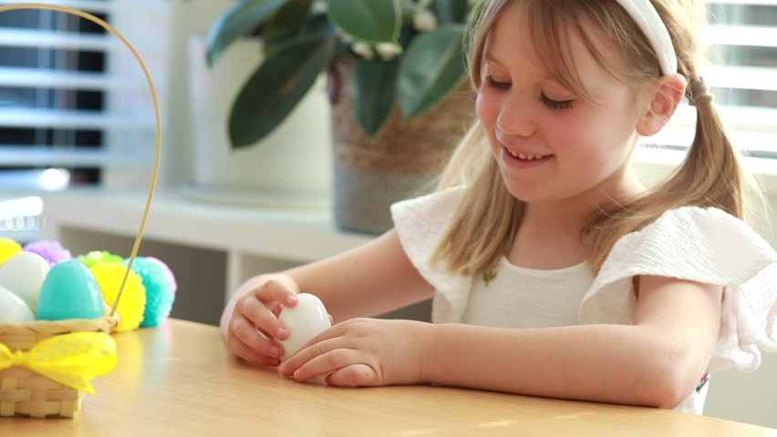 A cute little girl opens a plastic Easter egg with colorful jelly beans inside sitting at table. Concept of kids game egg hunt. Happy Easter.