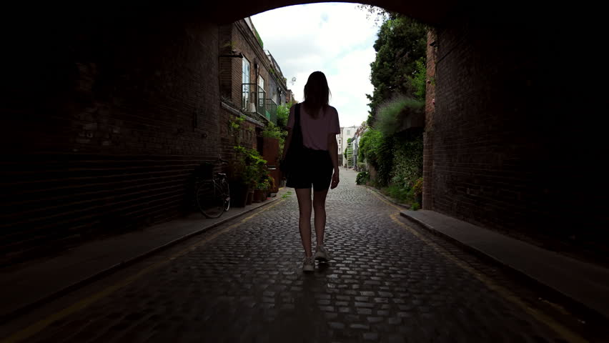 Follow footage rear view of silhouette of unrecognizable young woman in shorts walking forward in archway in old part of town. tourist traveling in city.