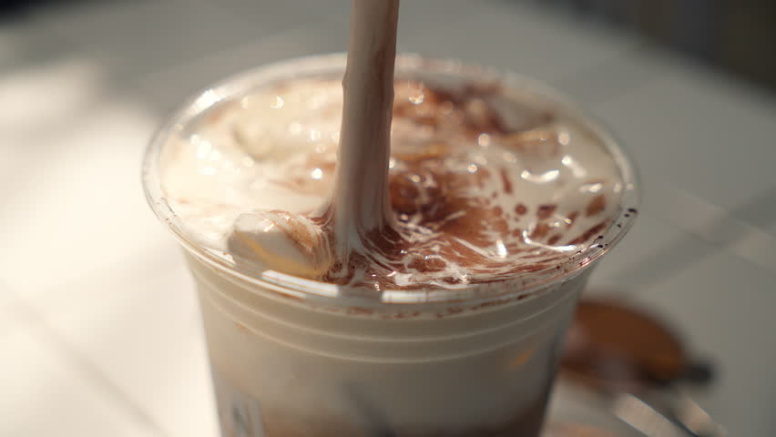 Close up view of iced coffee with cream and caramel, stirred with a drinking straw in a disposable eco-friendly cup.