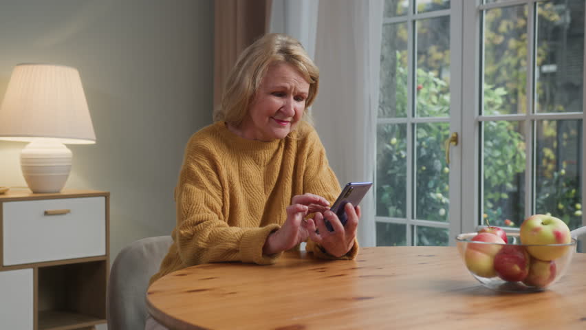 Elderly cheerful woman sits at table holding mobile phone. Senior woman smiles scrolling, enjoying typing. Female retired using cell phone for app online, looking through photos and videos