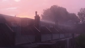 A row of houses with chimneys is seen under a pink-hued sky during rainfall. Raindrops blur the view, and trees in the background add depth to the misty atmosphere. The sunset casts a soft glow. - Powered by Shutterstock - Get 15% off with code: PIKWIZARD15