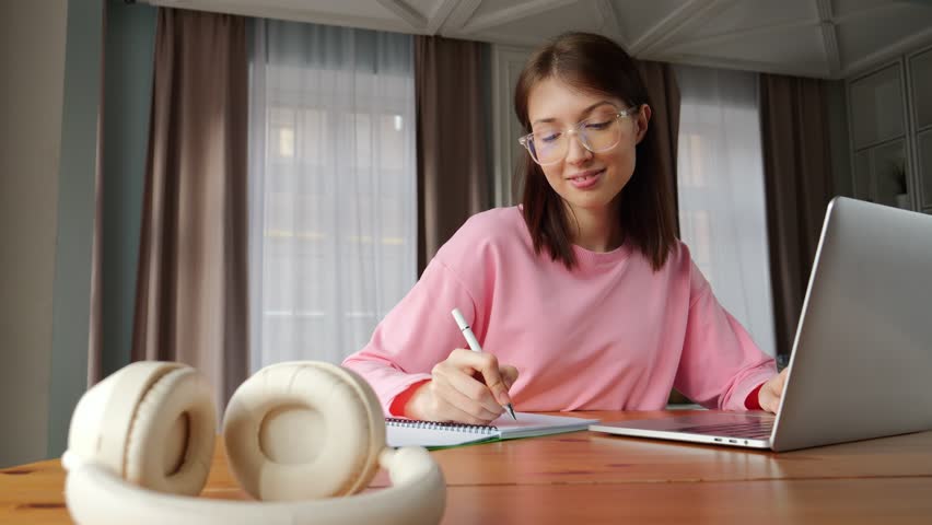 Woman writes notes while using her laptop at a wooden table in a warm, inviting living space.
