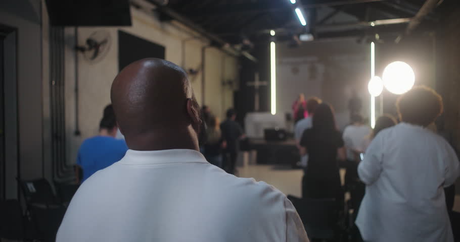 Wide shot of church service with congregation standing and worshipping, back view of people focused on stage, modern church interior, unity in faith