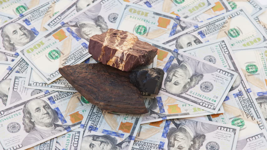 Three gemstones, Tiger Iron, Hematite, and Obsidian, rotate on a turntable atop genuine US 100-dollar bills. Dark stones with metallic and earthy tones contrast with the green currency background.