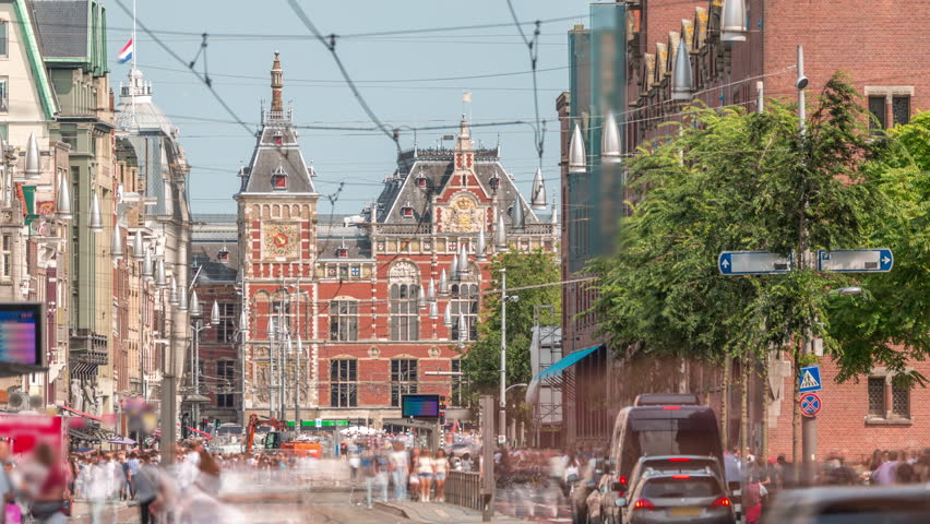 Damrak Street heading from Dam Square to Centraal Station in a timelapse with electric tram. Traffic moves on the road while people wait at the tram stop. Amsterdam, Netherlands