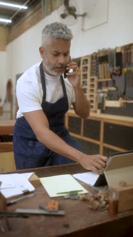 Small business owner wearing apron talking on phone, writing notes on clipboard and using tablet in woodworking workshop. Grey-haired carpenter multitasking in busy workshop