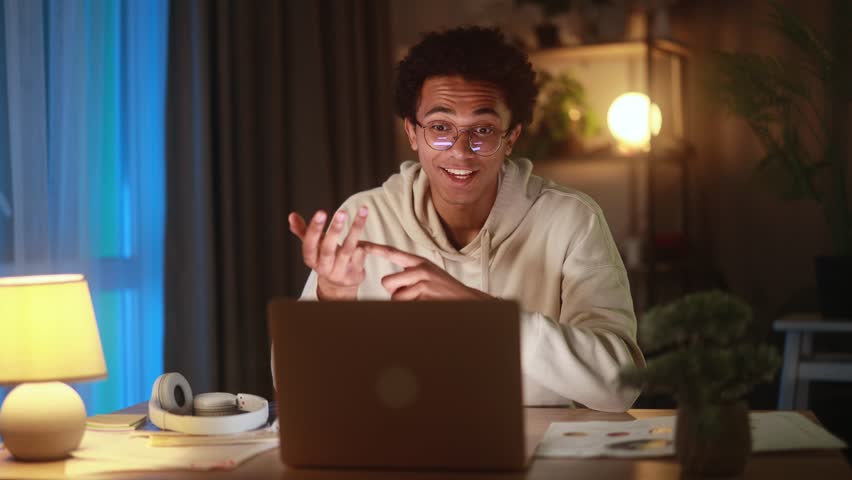 Cheerful young African American man wearing glasses and hoodie having a virtual video call at home, smiling and gesturing while talking to a friend or colleague in a cozy evening environment
