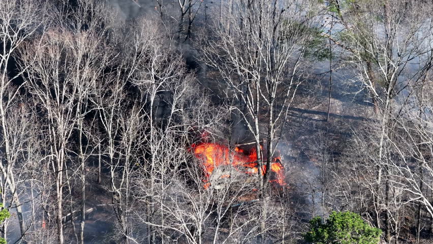 Telephoto aerial drone footage of a camper van that is on fire in the forest.
