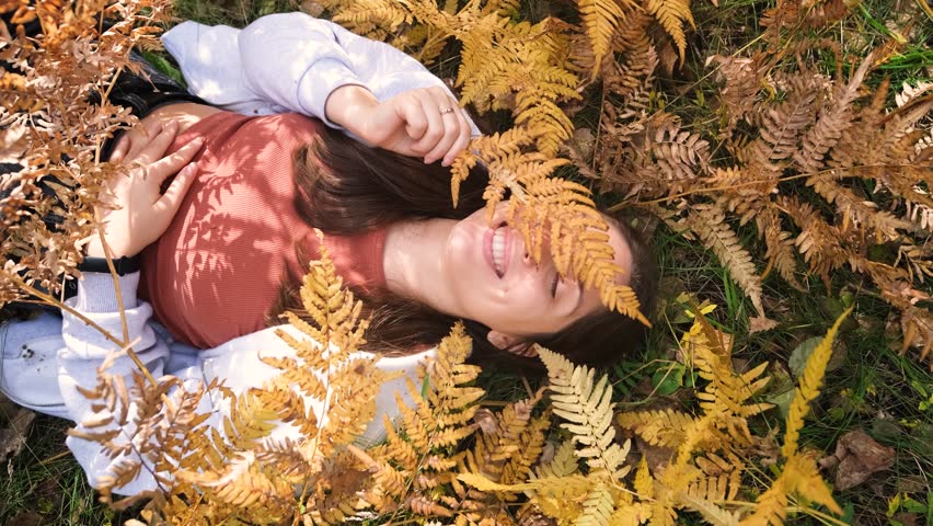 Cheerful young woman lies on yellow autumn leaves in the park and smiles.