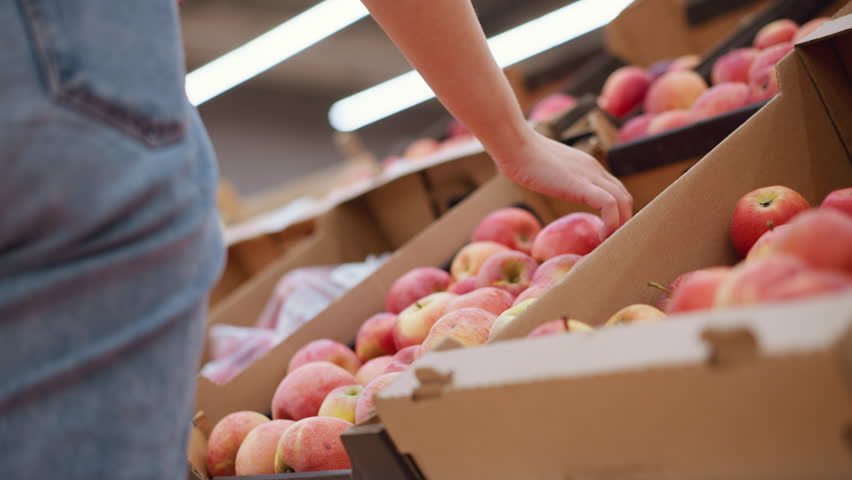 Back partial view of lady with phone in back pocket picking red apple from apples, observing it, and dropping it back in a vibrant grocery store setting