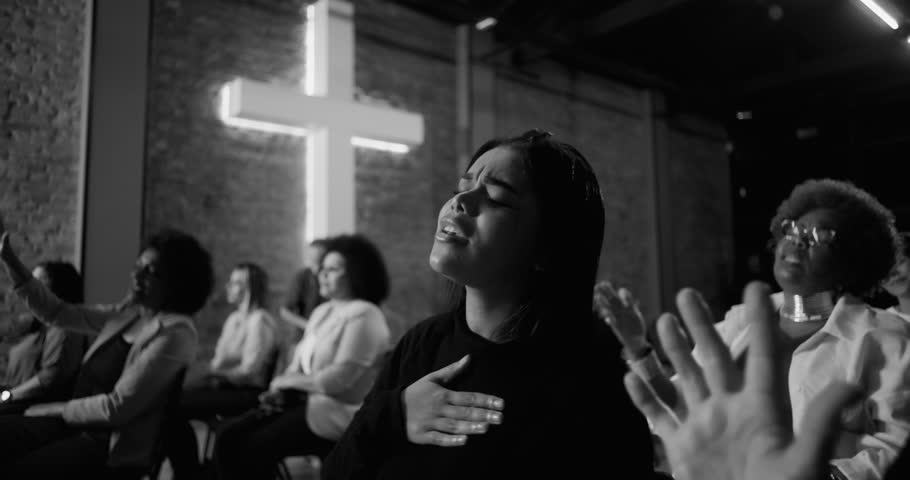 Young woman singing in prayer during worship service, hand on chest in devotion, illuminated cross and congregation in background, black and white tones