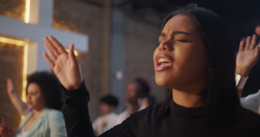 Young woman singing passionately in worship, raising hand in church surrounded by congregation, heartfelt devotion and spiritual expression during prayer